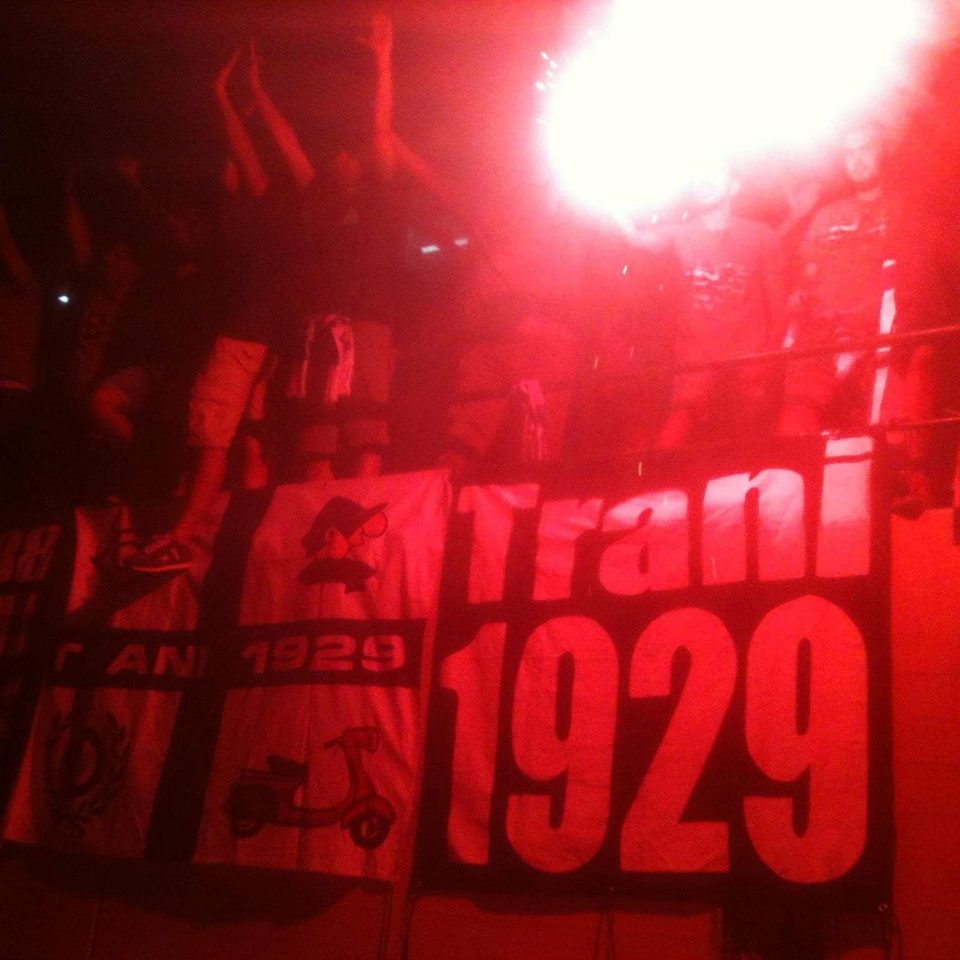 Striscione Trani 1929 illuminato dai fumogeni rossi in curva